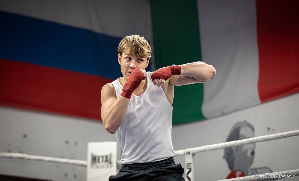 Un jeune boxeur en débardeur blanc, mains bandées de rouge, se tient en garde devant les cordes d’un ring